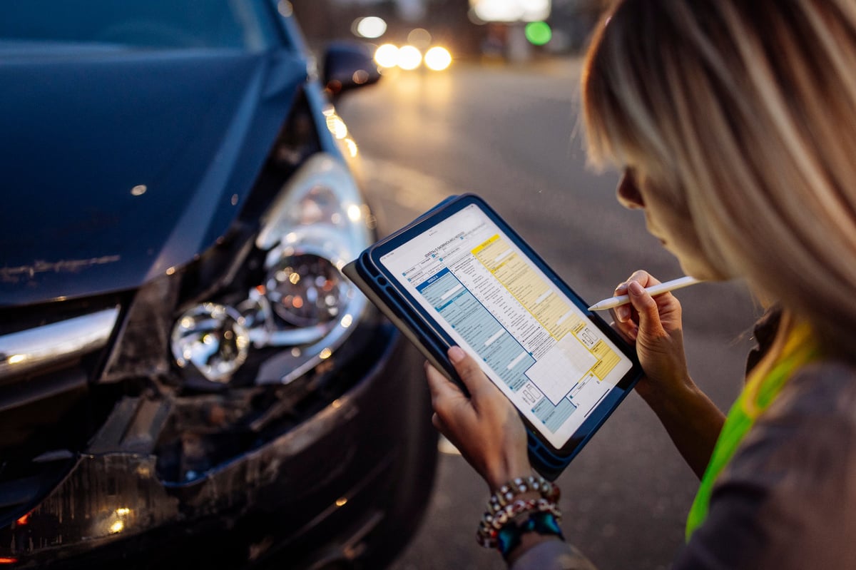 Young woman wearing a reflective safety vest writes on a clipboard or digital tablet near a damaged car after a traffic accident