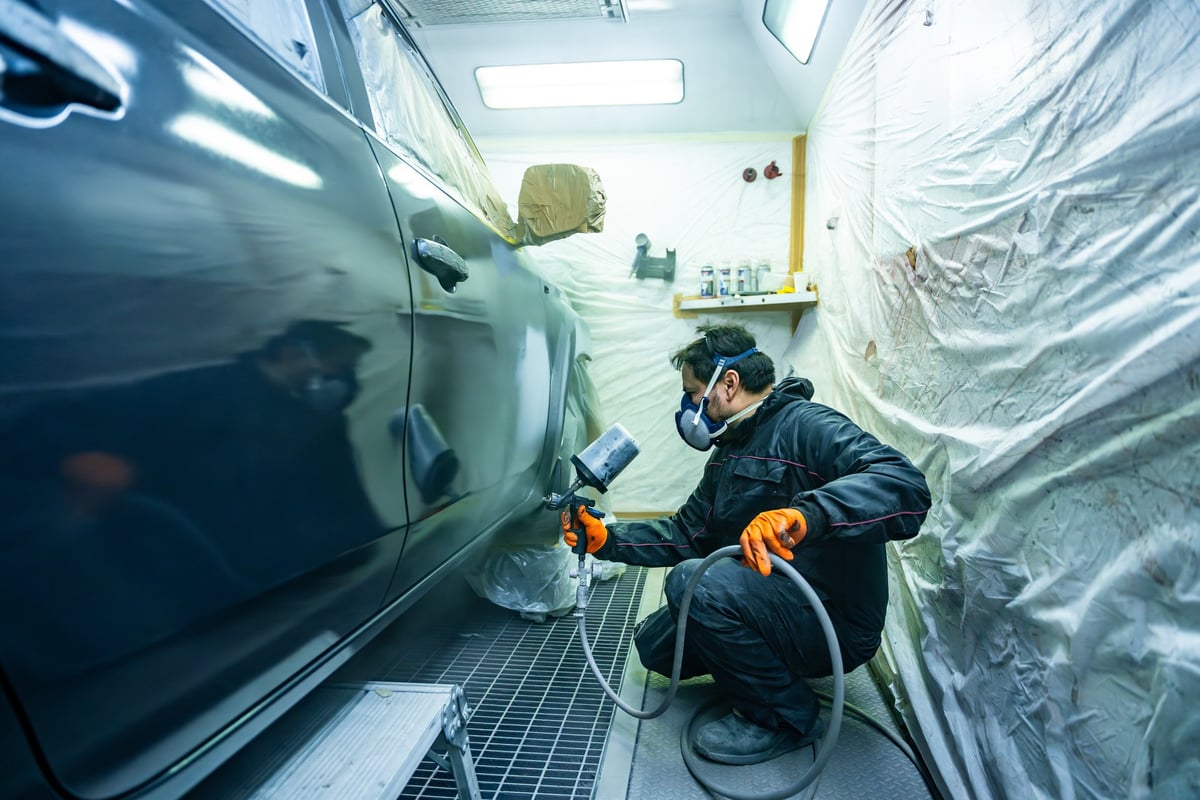 Automotive technician wearing a protective mask and gloves is carefully applying primer to a car panel in a specialized paint spray booth at an auto body repair shop